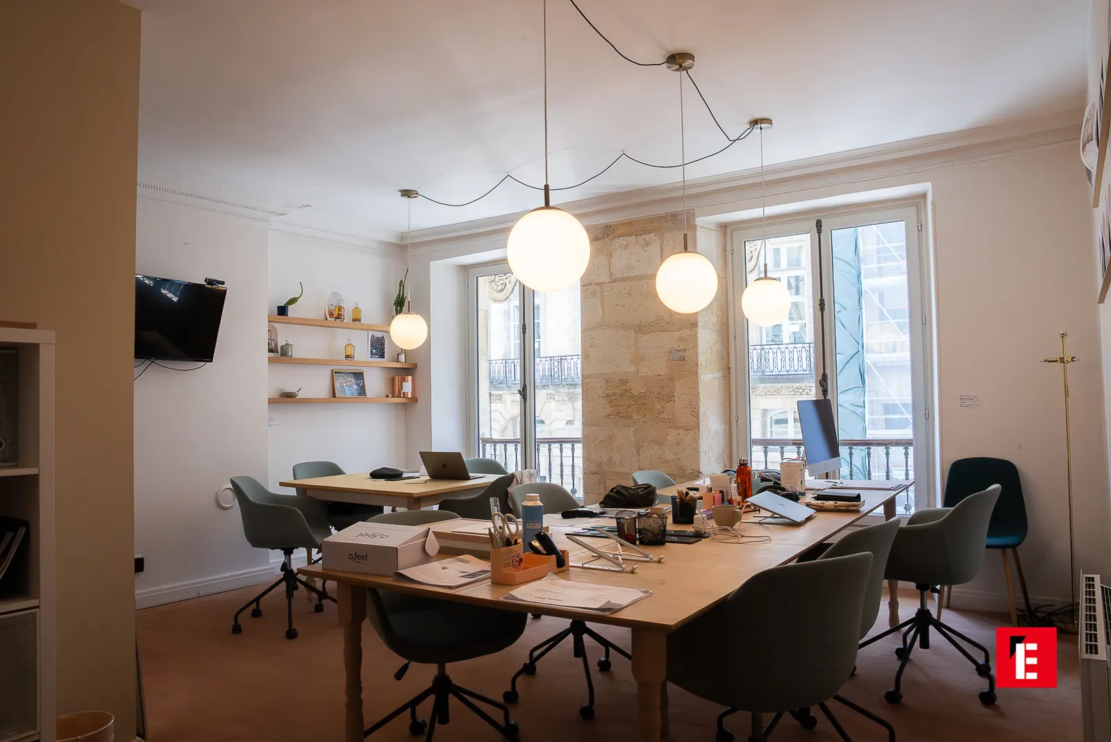 Grande salle de coworking aux suspensions globe, tables en bois et fauteuils verts face aux fenêtres sur la rue bordelaise — Bordeaux Esprit des Lois